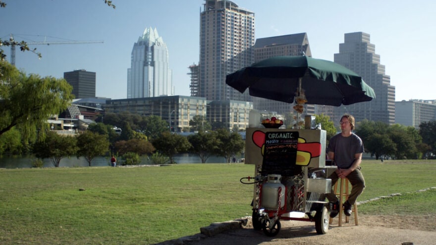 Bill sitting with a hotdog stand in auditorium shores park with Austin skyline in background