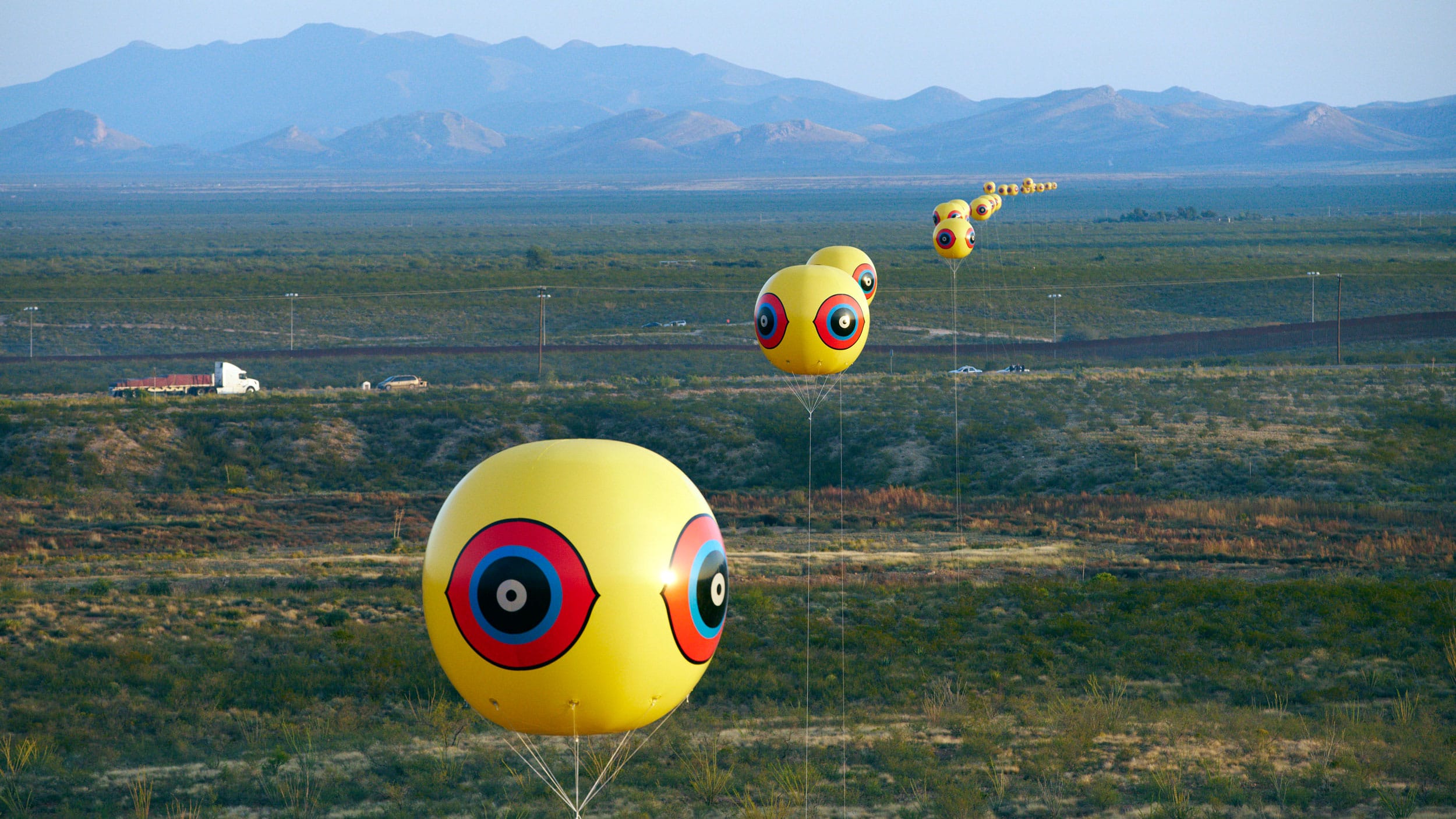 Giant yellow balloons with red and black designs in a row for miles through a landscape with mountains in the background